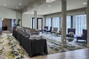 Uma longa mesa de buffet com talheres e pratos, rodeada por cadeiras e uma grande janela com cortinas.