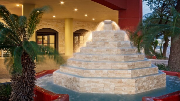Una fuente de agua con una escalera de piedra, palmeras y un edificio con puertas arqueadas.