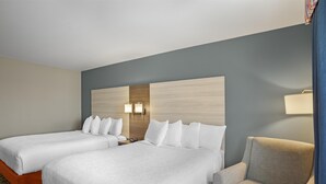 Ein Hotelzimmer mit zwei Betten, einem grauen Sessel und einem Holzkopfstück mit Wandleuchten.