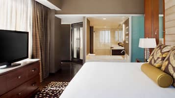 Habitación de hotel con una cama grande, un televisor sobre una cómoda de madera, un baño con espejo y lavamanos, y un pasillo que da a otra habitación.