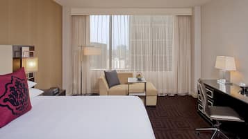 A hotel room with a large bed, a desk, a chair, a small table, and a window with sheer curtains.