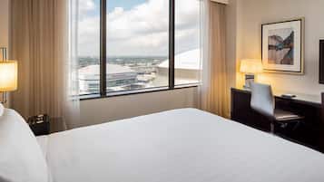 A hotel room with a large bed, a desk with a chair, a TV, and a view of a stadium through a large window.