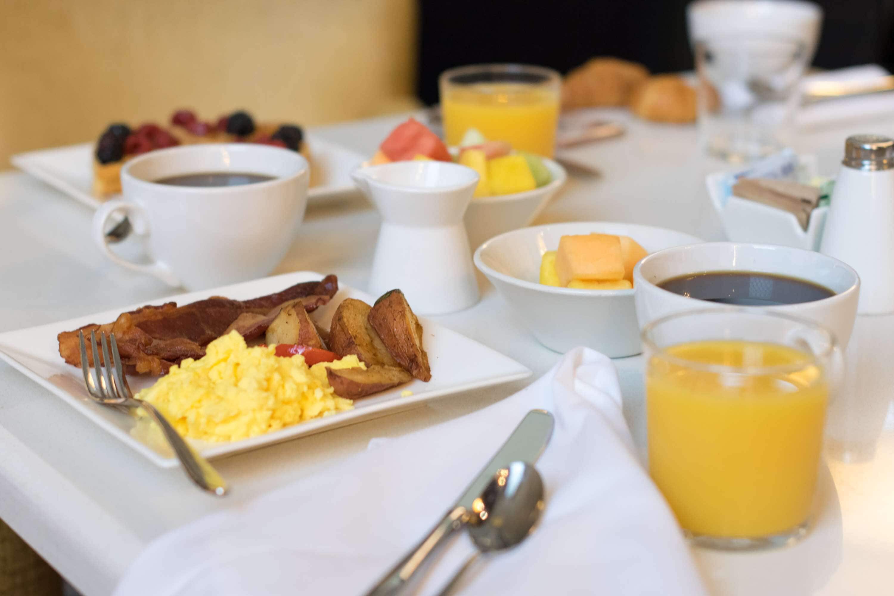 A breakfast table with plates of eggs, bacon, toast, and fruit. There are cups of coffee and orange juice.