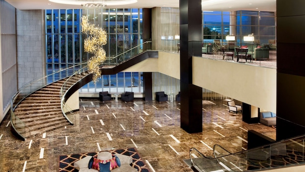 A spacious lobby with a curved staircase, a seating area with a patterned rug, and large windows offering a view of the cityscape.