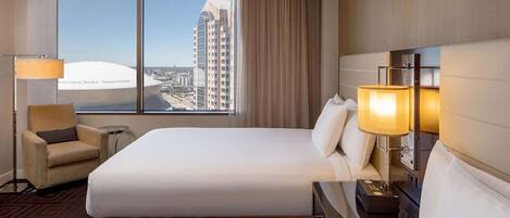 A hotel room with a large bed, a chair, a bedside table with a lamp, and a view of a cityscape.