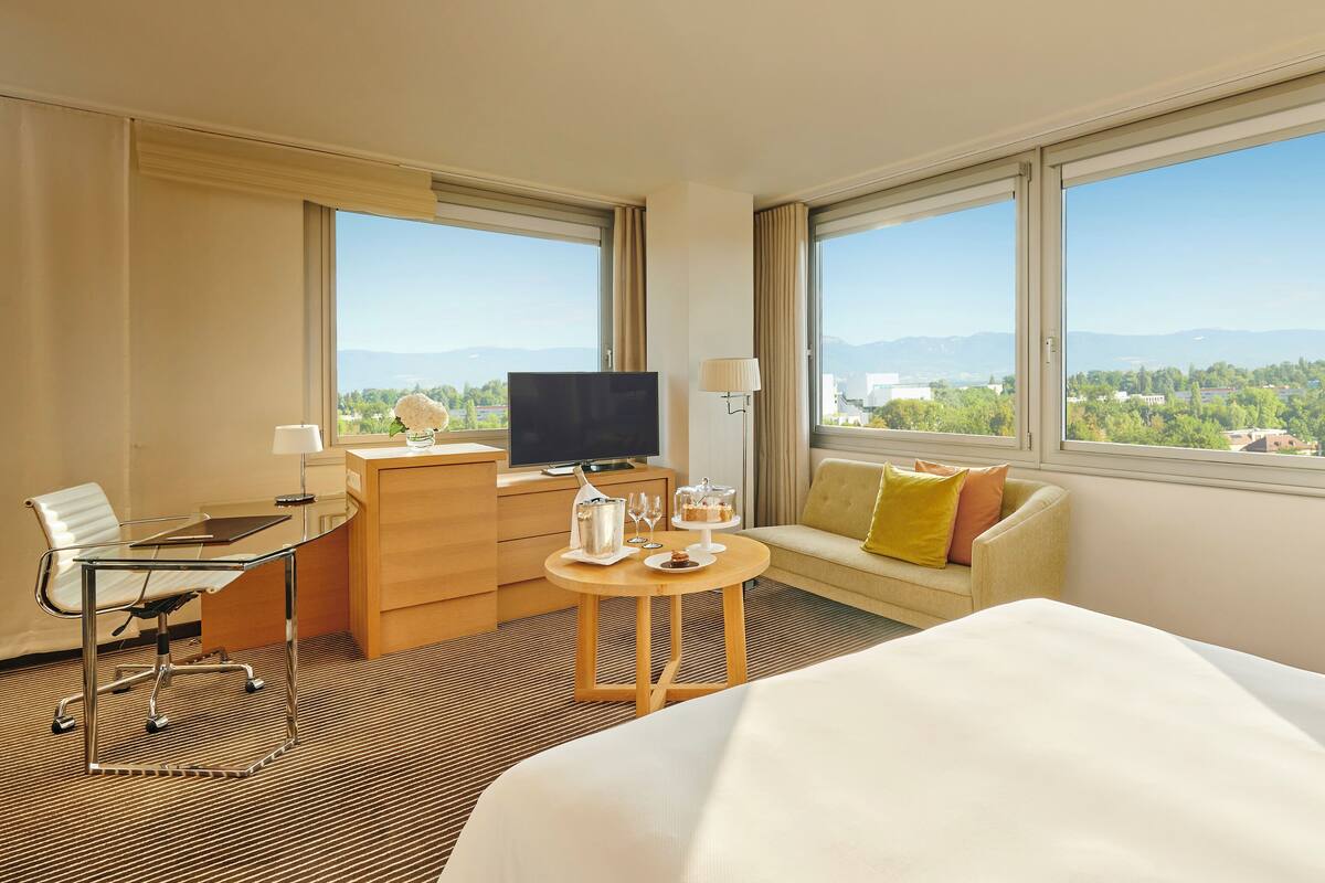 A hotel room with a bed, a desk with a computer, a TV, a sofa, and large windows with a city view.