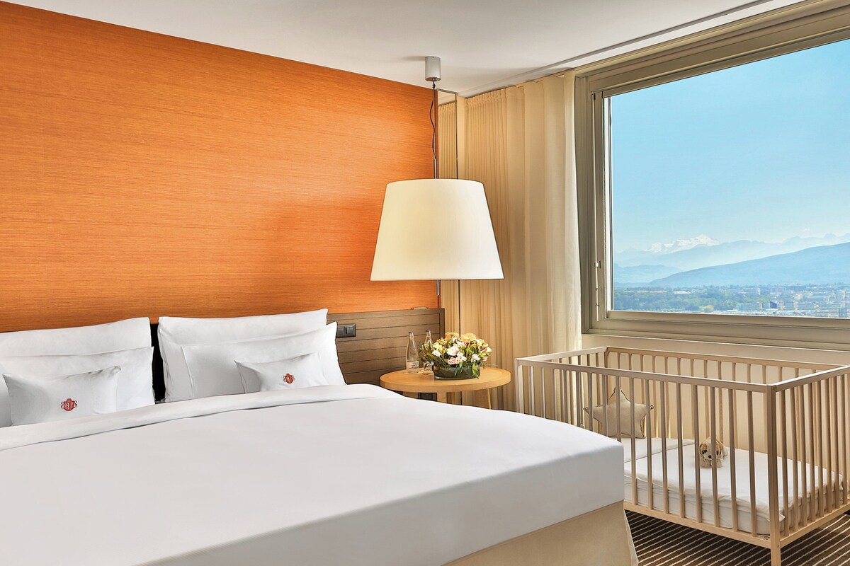 A hotel room with a bed, a crib, a nightstand with a lamp, and a window with a city and mountain view.