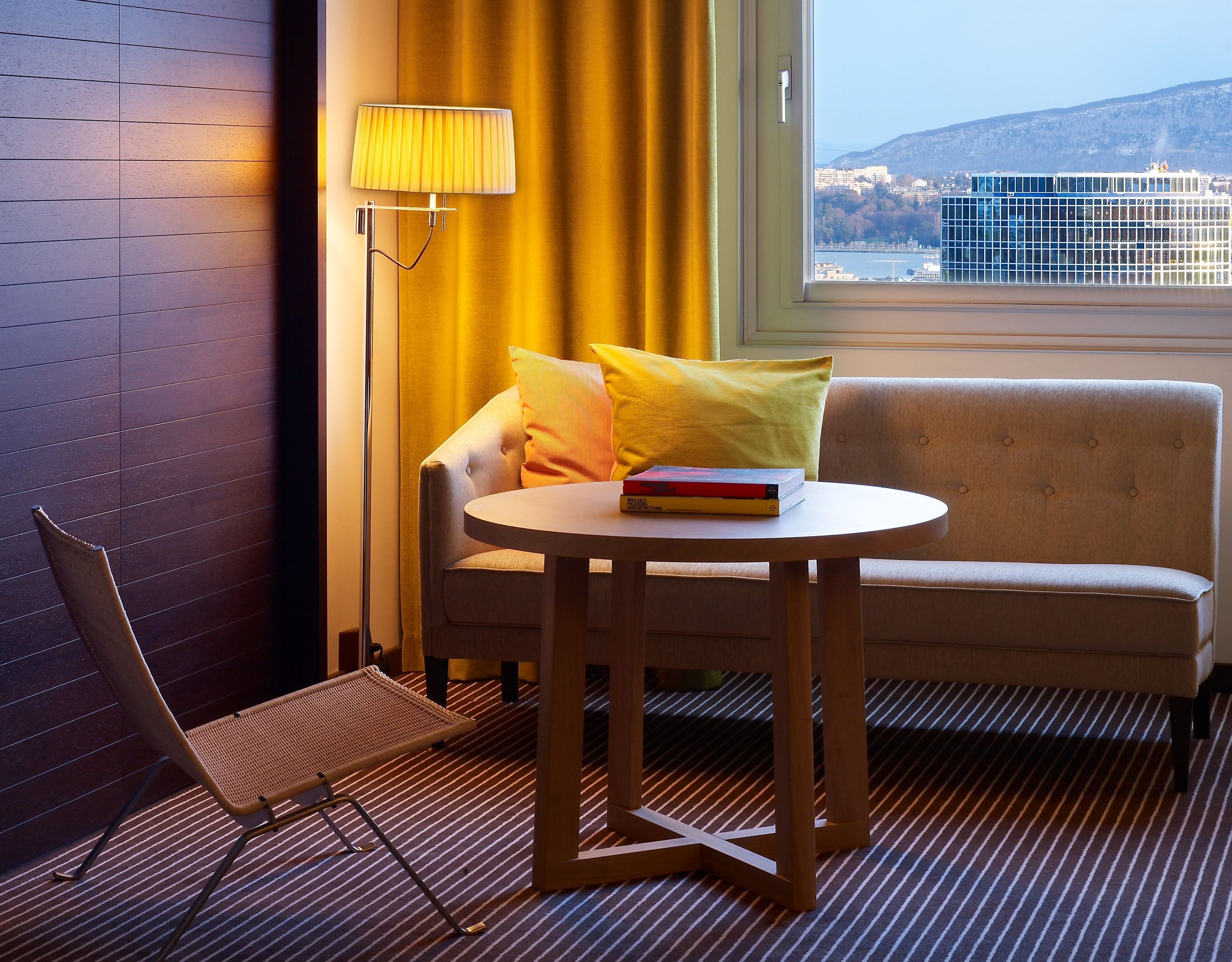 A hotel room with a sofa, a chair, a small table, a lamp, and a view of a cityscape through a window.