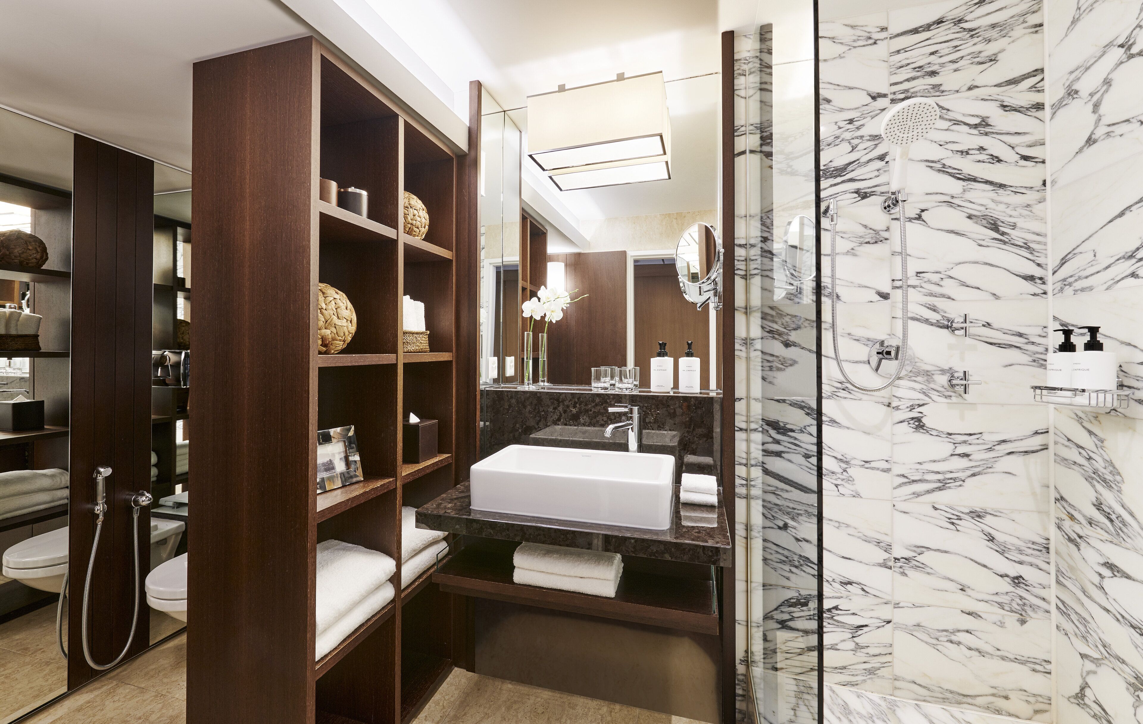 A modern bathroom with a large mirror, a white sink, and a shower area. There are shelves with toiletries and towels.