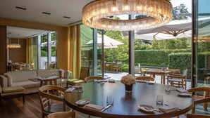 A dining area with a round table set for four, surrounded by wooden chairs. A large, circular chandelier hangs above the table. The room features large windows offering a view of a garden and outdoor seating area.