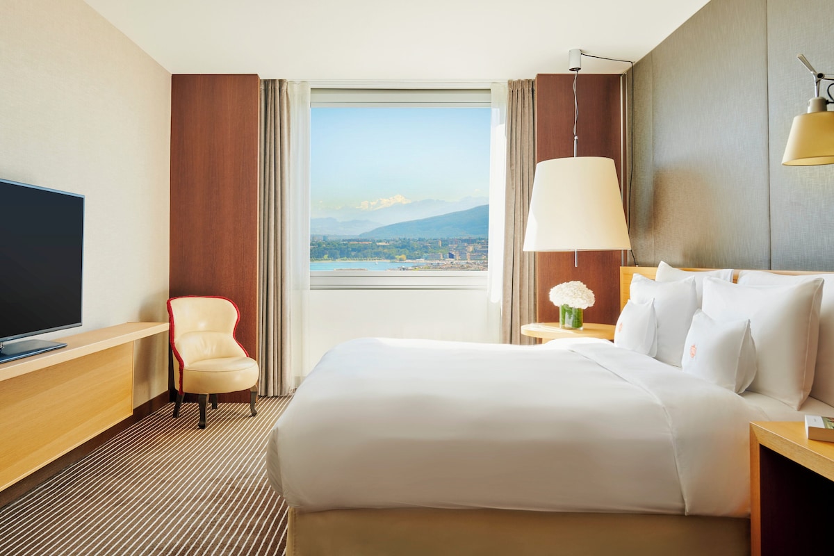 Habitación de hotel con una cama grande, televisión, silla, lámpara y vistas a un lago y montañas.