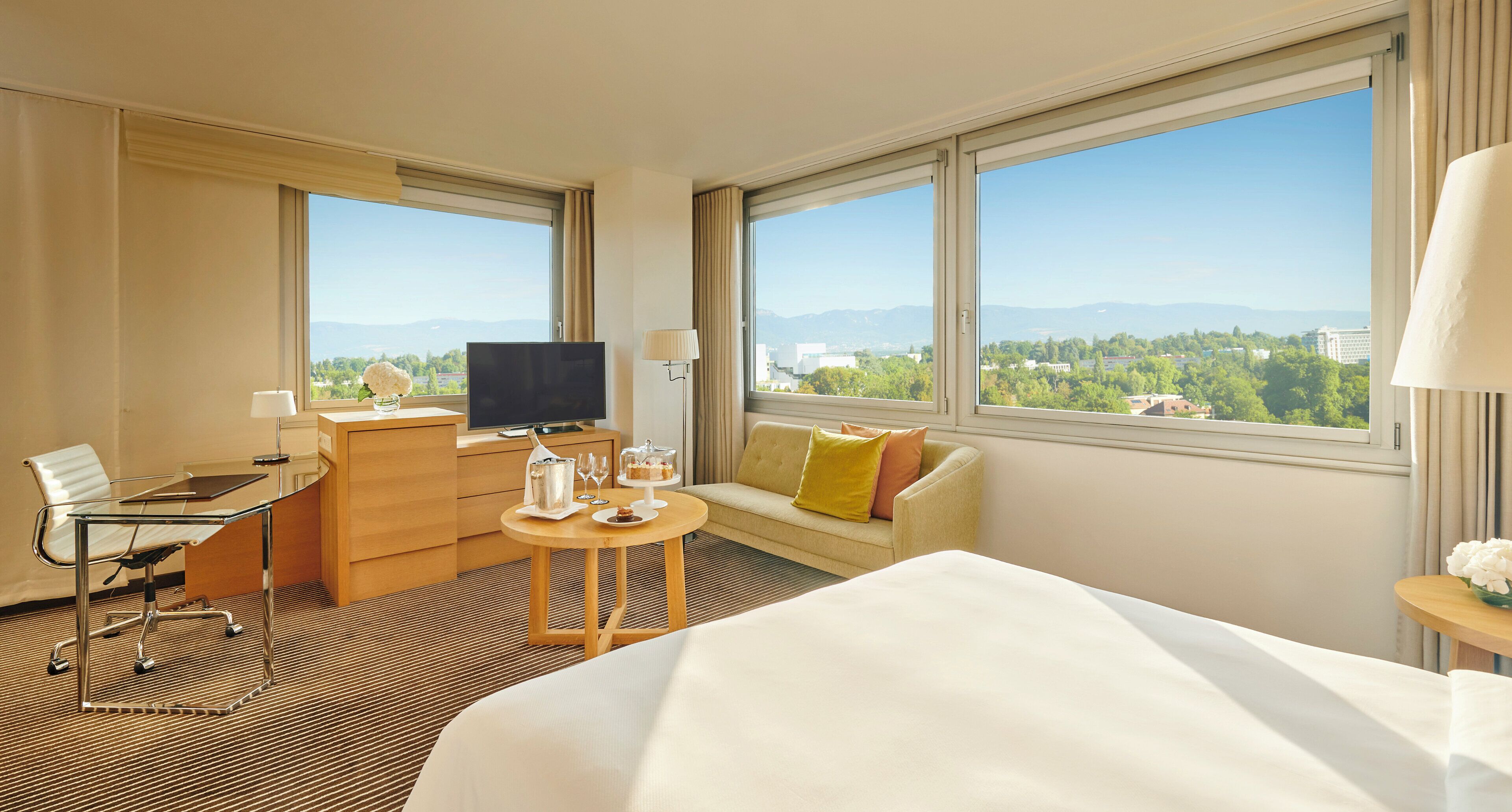 A hotel room with a bed, a desk with a chair, a sofa, a television, and large windows offering a city view.