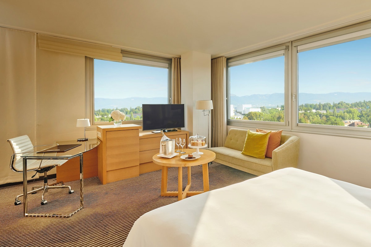 Habitación de hotel con cama, escritorio con silla, sofá, televisión y ventanas grandes con vistas a la ciudad.