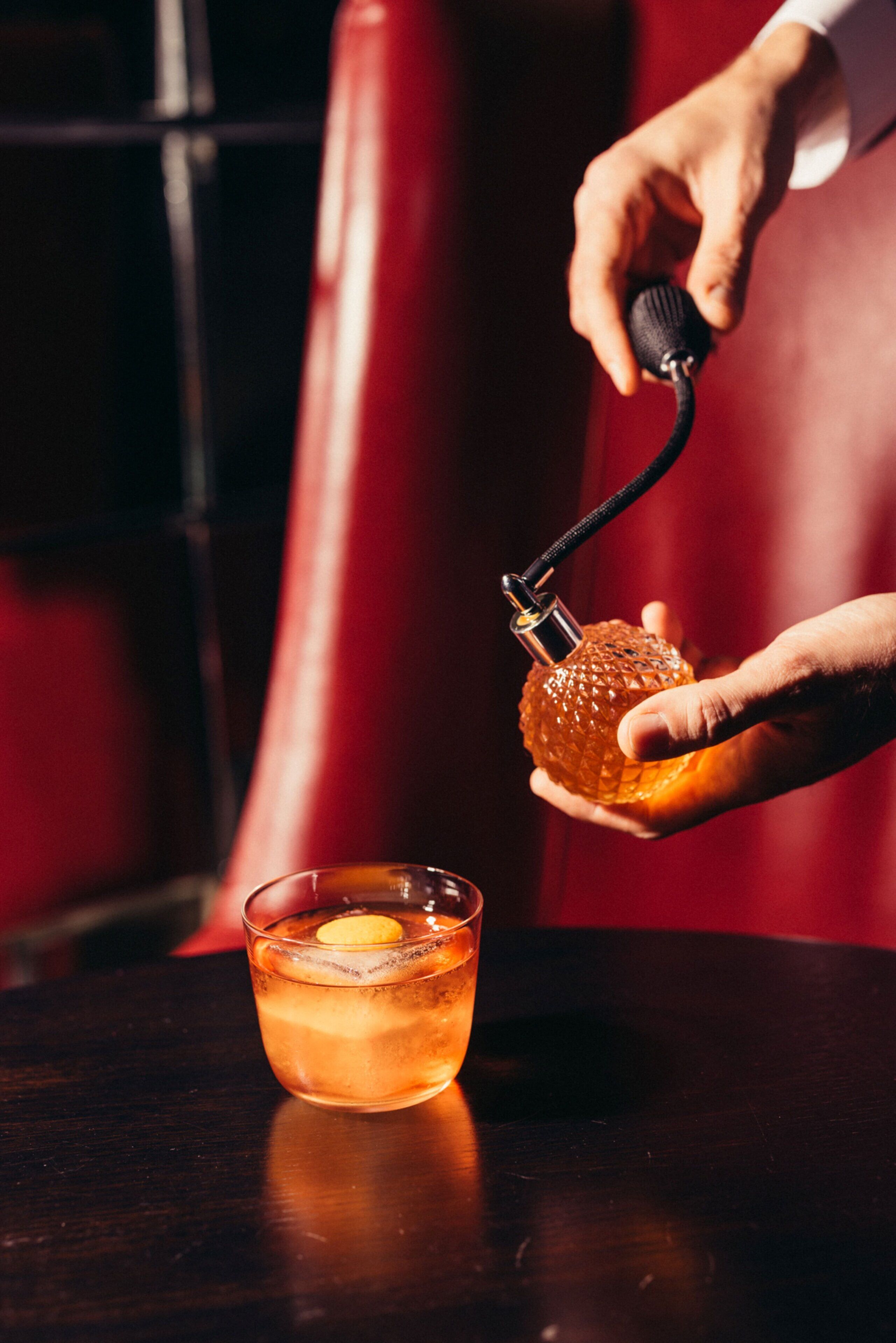 A hand holding a cocktail shaker with a lemon twist, pouring into a glass with ice.