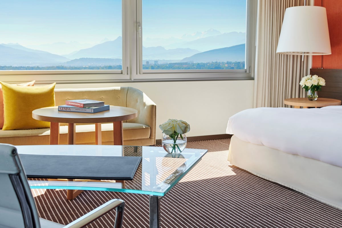 Habitación de hotel con cama, sofá, una mesa redonda y una silla. Se ve una ventana con vistas a las montañas.