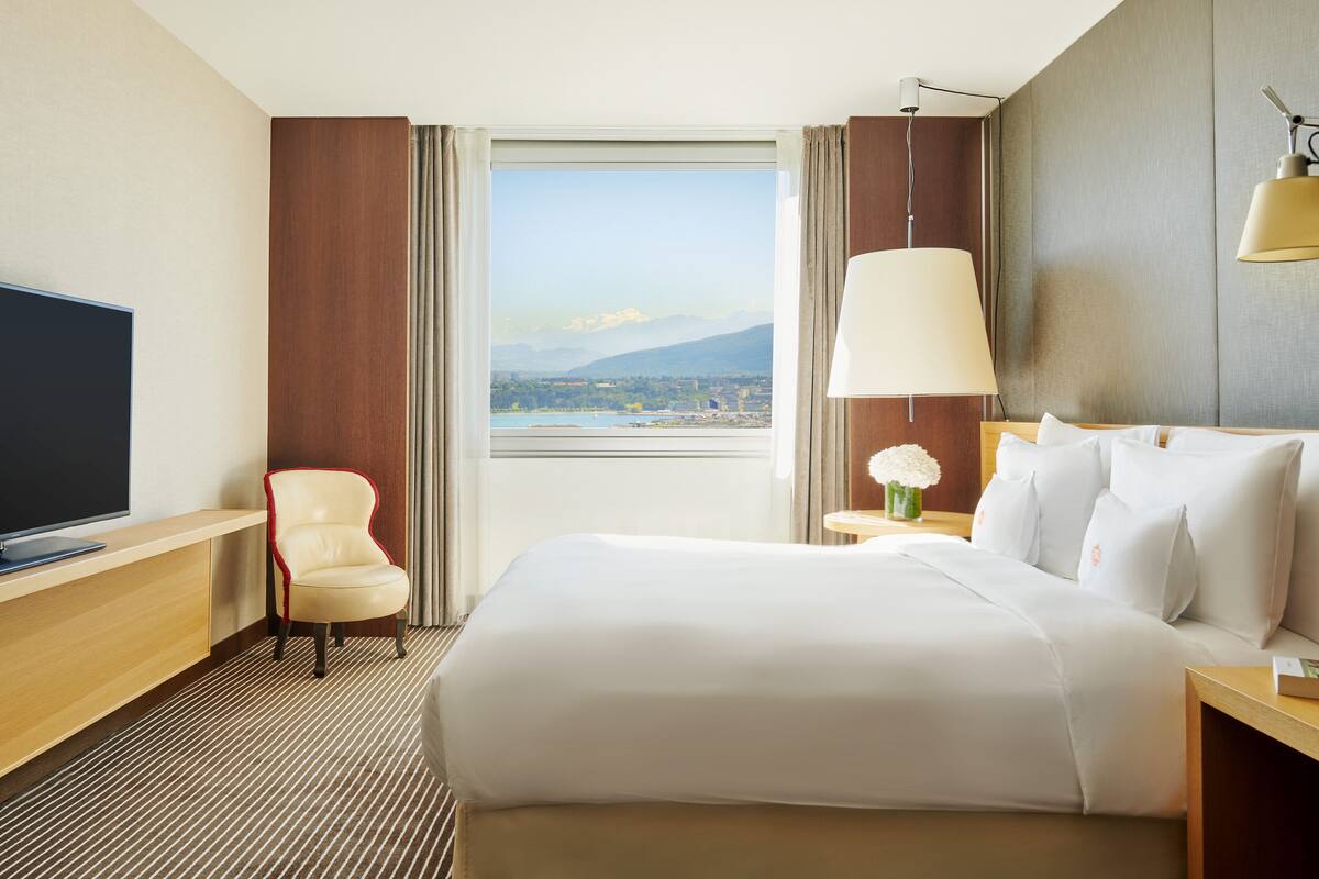 A hotel room with a large bed, a TV, a chair, a lamp, and a view of a lake and mountains.