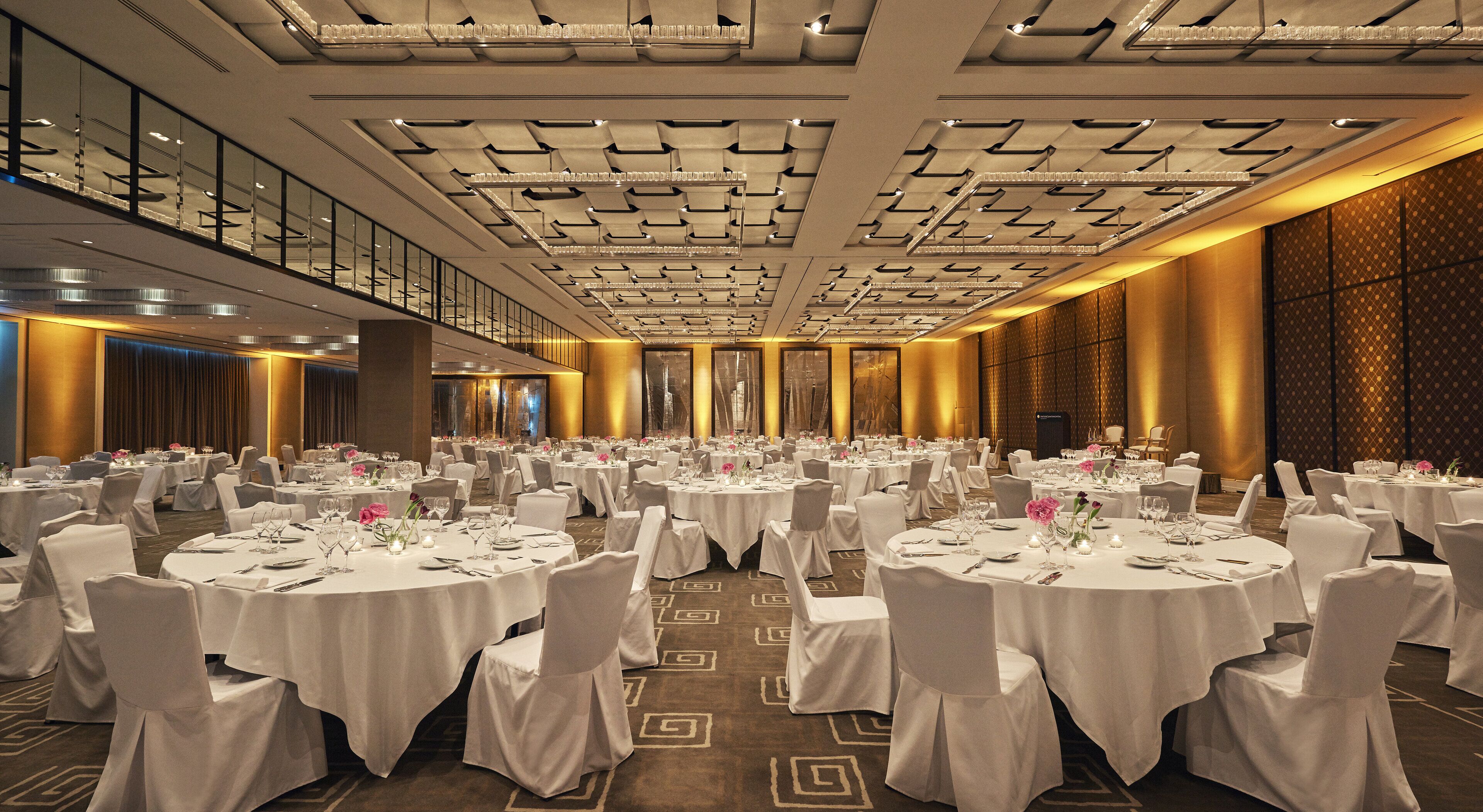 Une salle de banquet avec des tables rondes, des nappes blanches et des chaises, dotée d’un plafond haut avec des luminaires encastrés et d’un tapis à motifs.