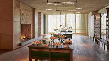 Un intérieur moderne avec une longue table à manger dressée et ornée de divers desserts, de grandes fenêtres équipées de stores et une cheminée.