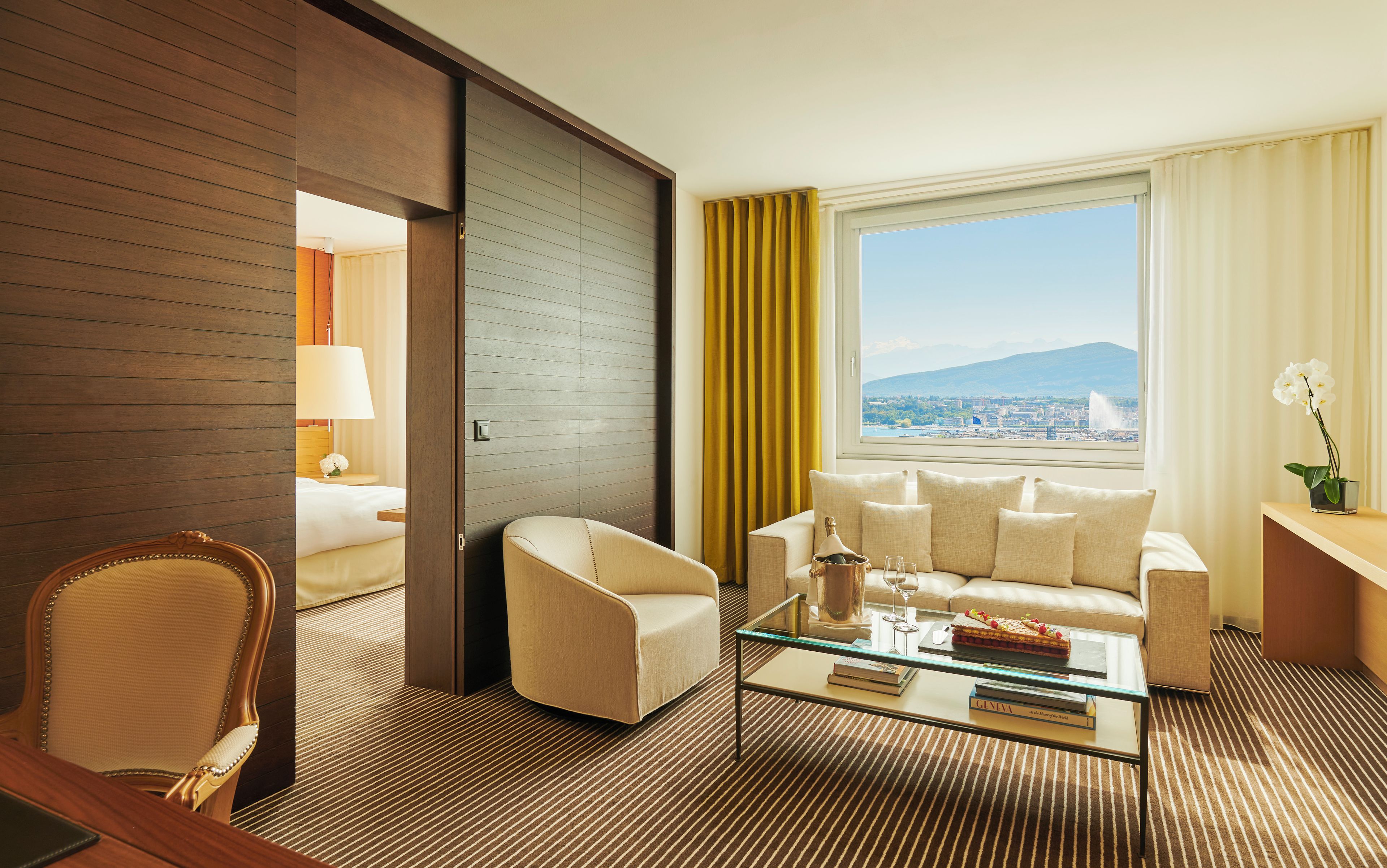 Une chambre d’hôtel avec un canapé, des fauteuils, une table basse en verre et une vue sur le paysage urbain et les montagnes.