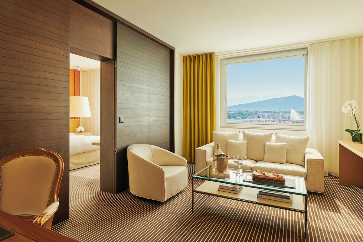 A hotel room with a sofa, armchairs, a glass coffee table, and a view of a cityscape and mountains.