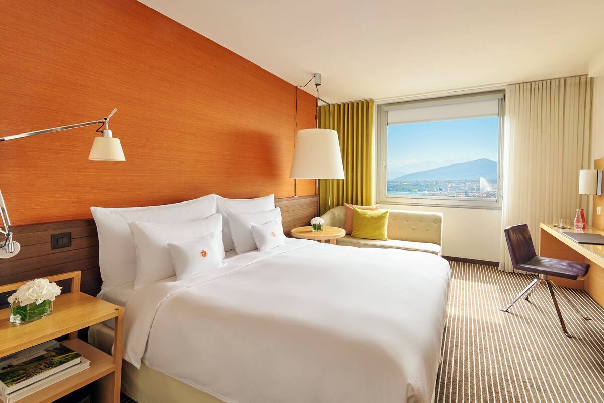 A hotel room with a large bed, a desk, a chair, a lamp, a window with a view, and a television.