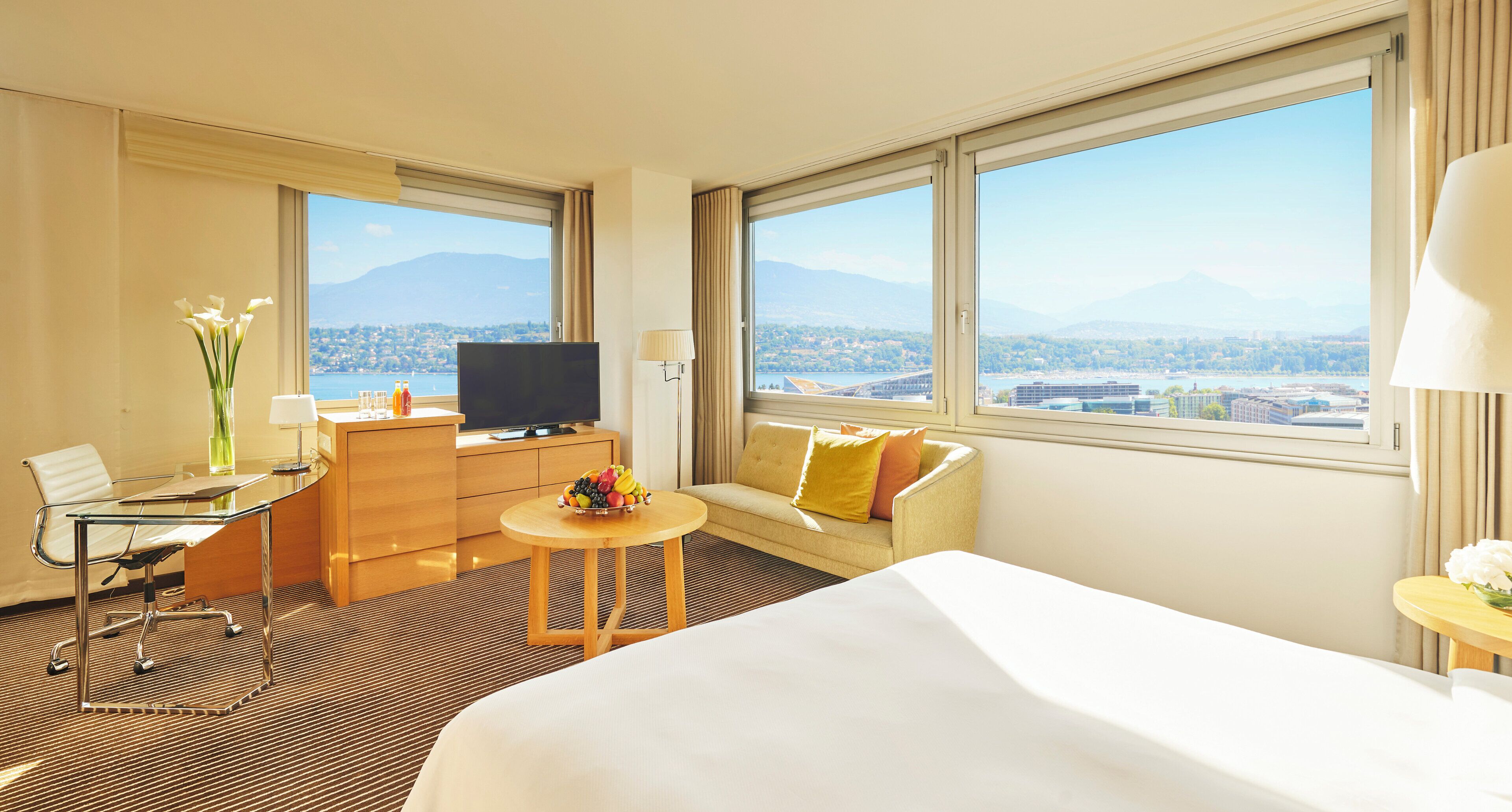 A hotel room with a bed, a desk with a computer, a television, a sofa, and a view of a cityscape.
