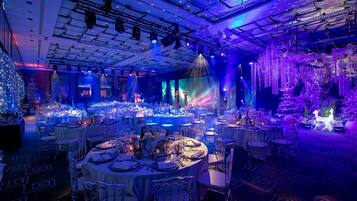 Un salón de banquetes con mesas redondas dispuestas para un evento formal, con un techo que tiene lámparas y un fondo con una proyección.