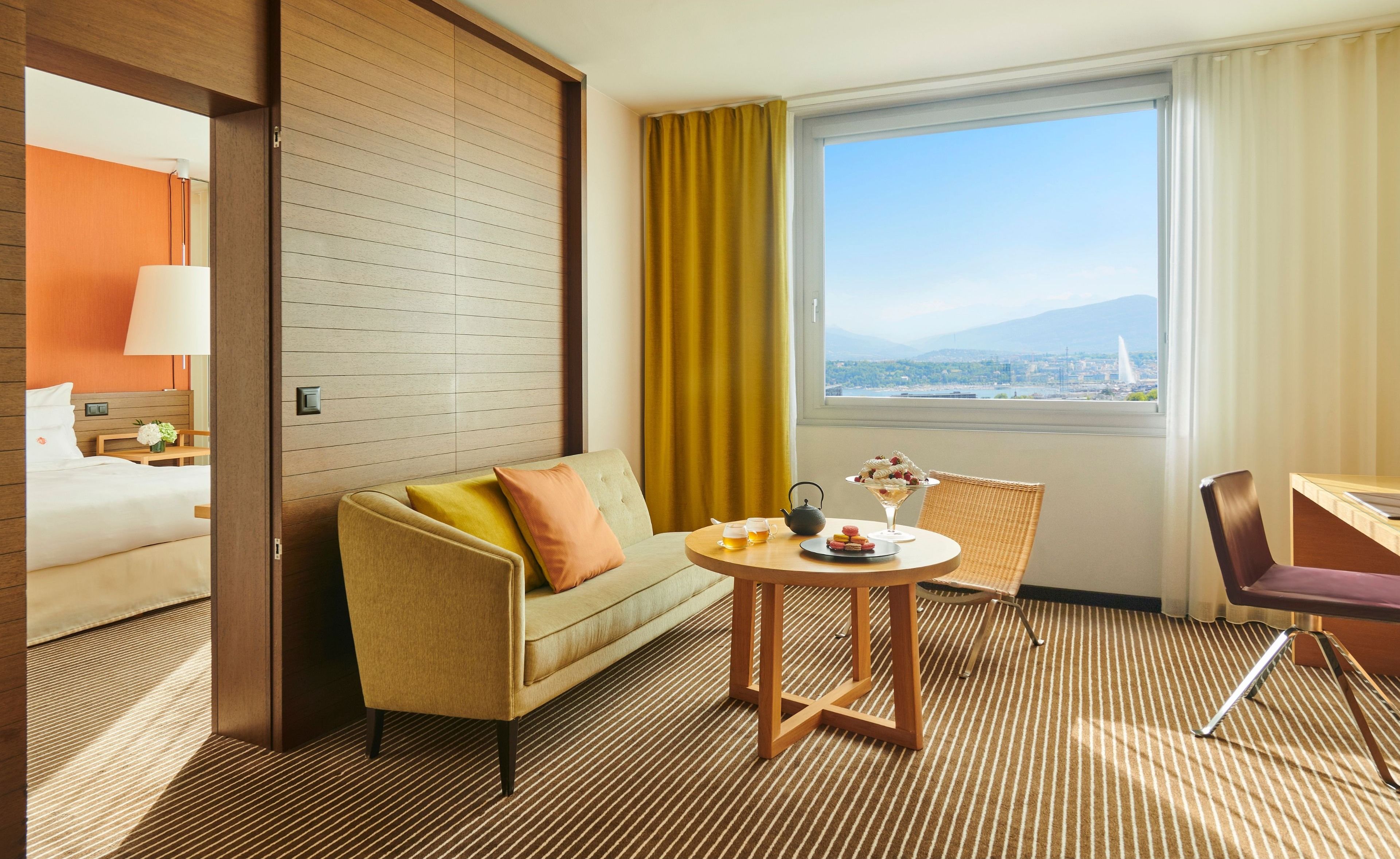 Habitación de hotel con sofá, una mesita redonda con tetera y tazas, una silla, una cama, una ventana con vistas y una lámpara.