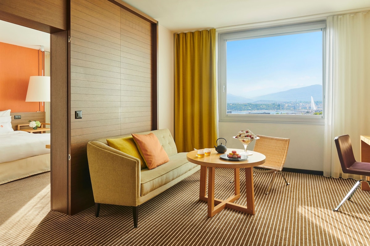 A hotel room with a sofa, a small round table with a teapot and cups, a chair, a bed, a window with a view, and a lamp.