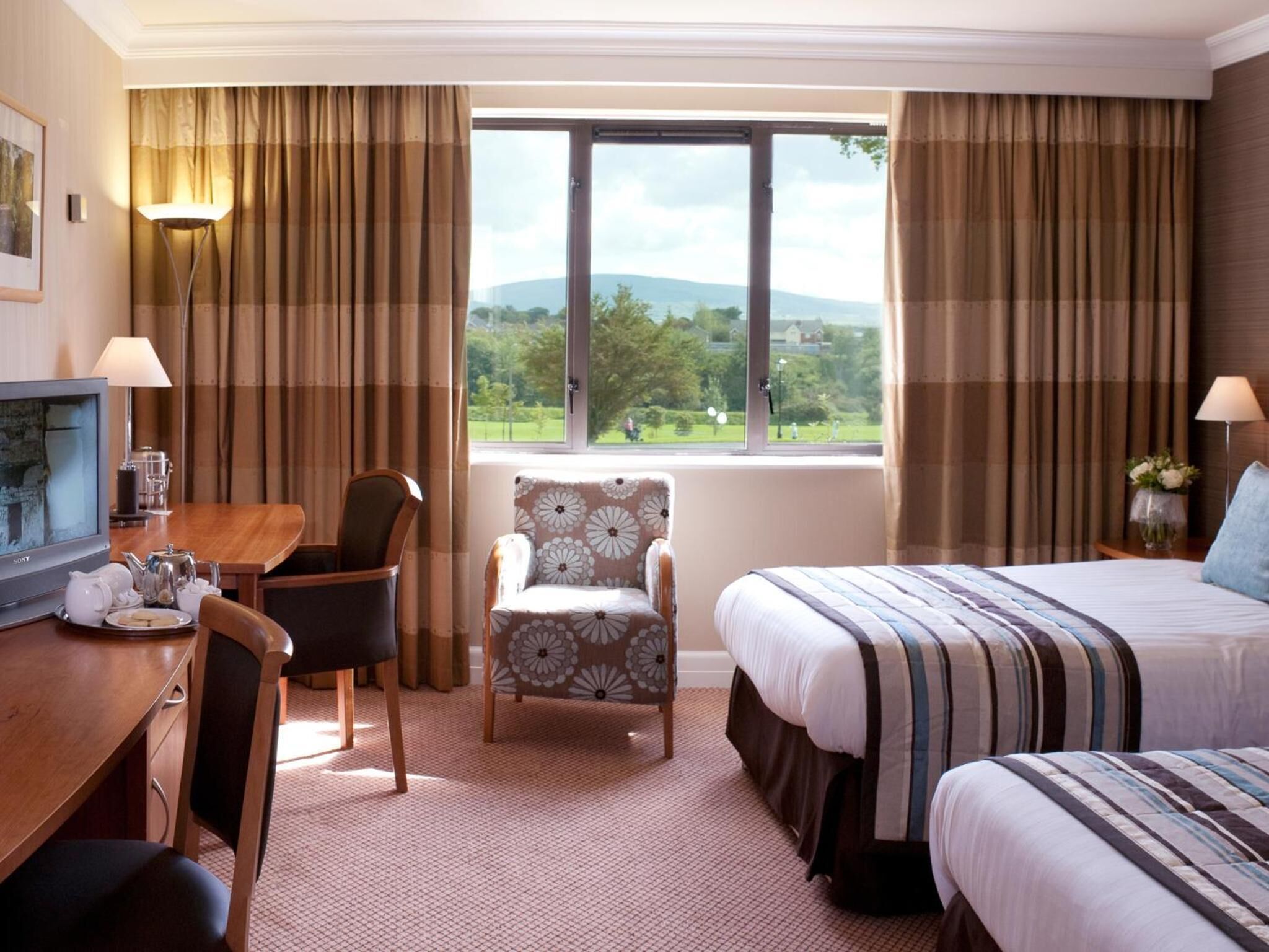 A hotel room with a bed, a chair, a desk with a television, and a window with a view of greenery and hills.