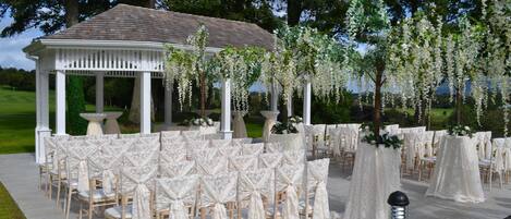 A gazebo with white columns and a tiled roof, surrounded by rows of chairs with white covers, set in a grassy outdoor area.