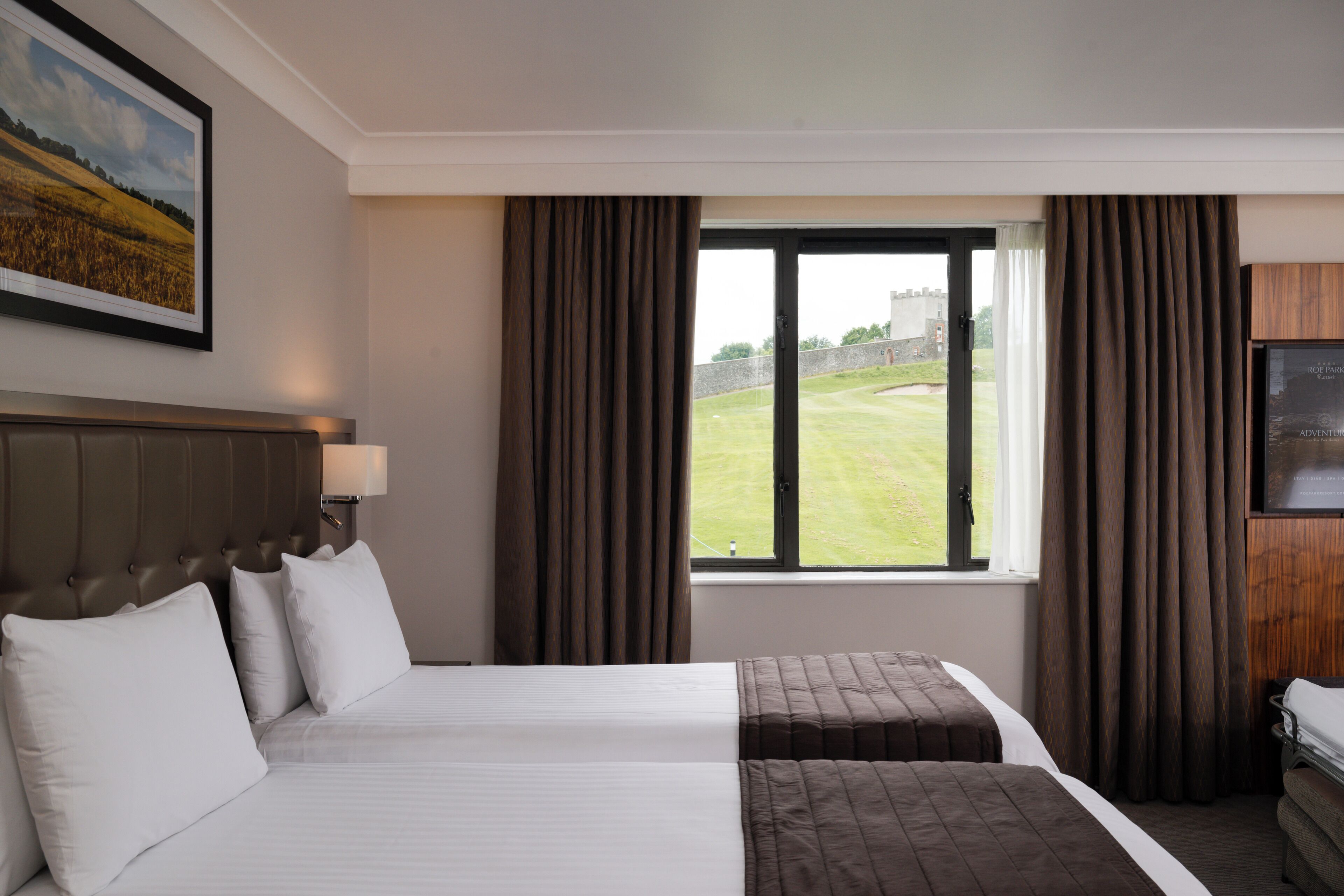 A hotel room with two beds, a large window with curtains, a mounted TV, and a framed picture on the wall.