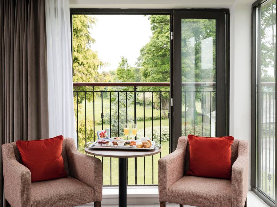A balcony with two armchairs, a table with breakfast items, and a view of greenery.