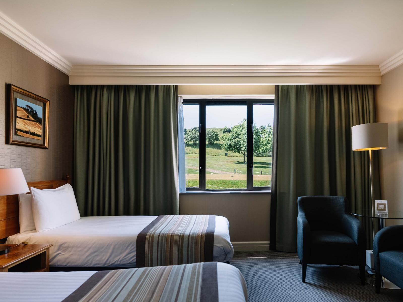 A hotel room with two beds, a desk, a chair, a lamp, and a large window with curtains.