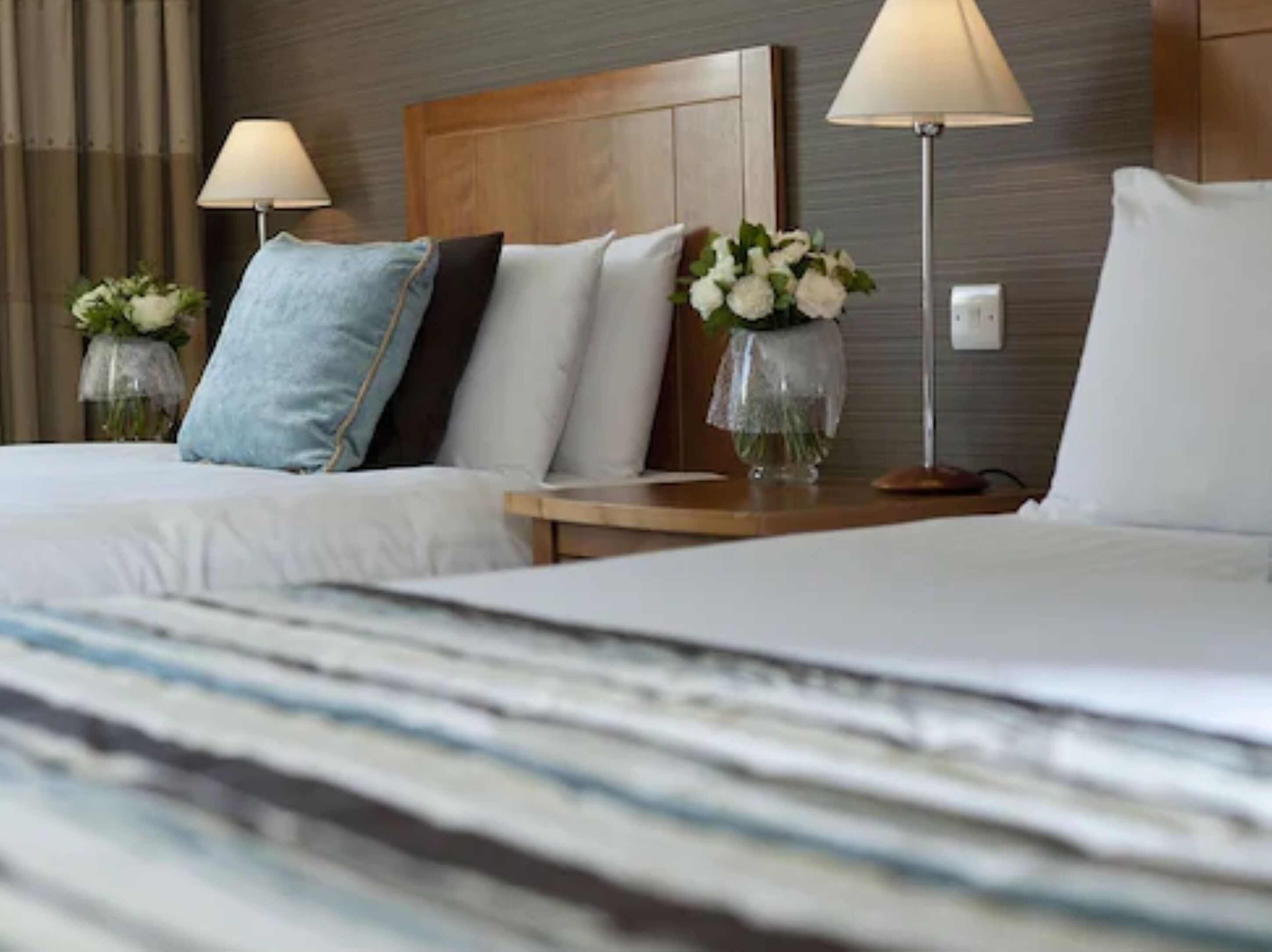 Ein Hotelzimmer mit zwei Betten, jedes mit einem Kissen, einer Vase mit Blumen und einer Nachttischlampe.