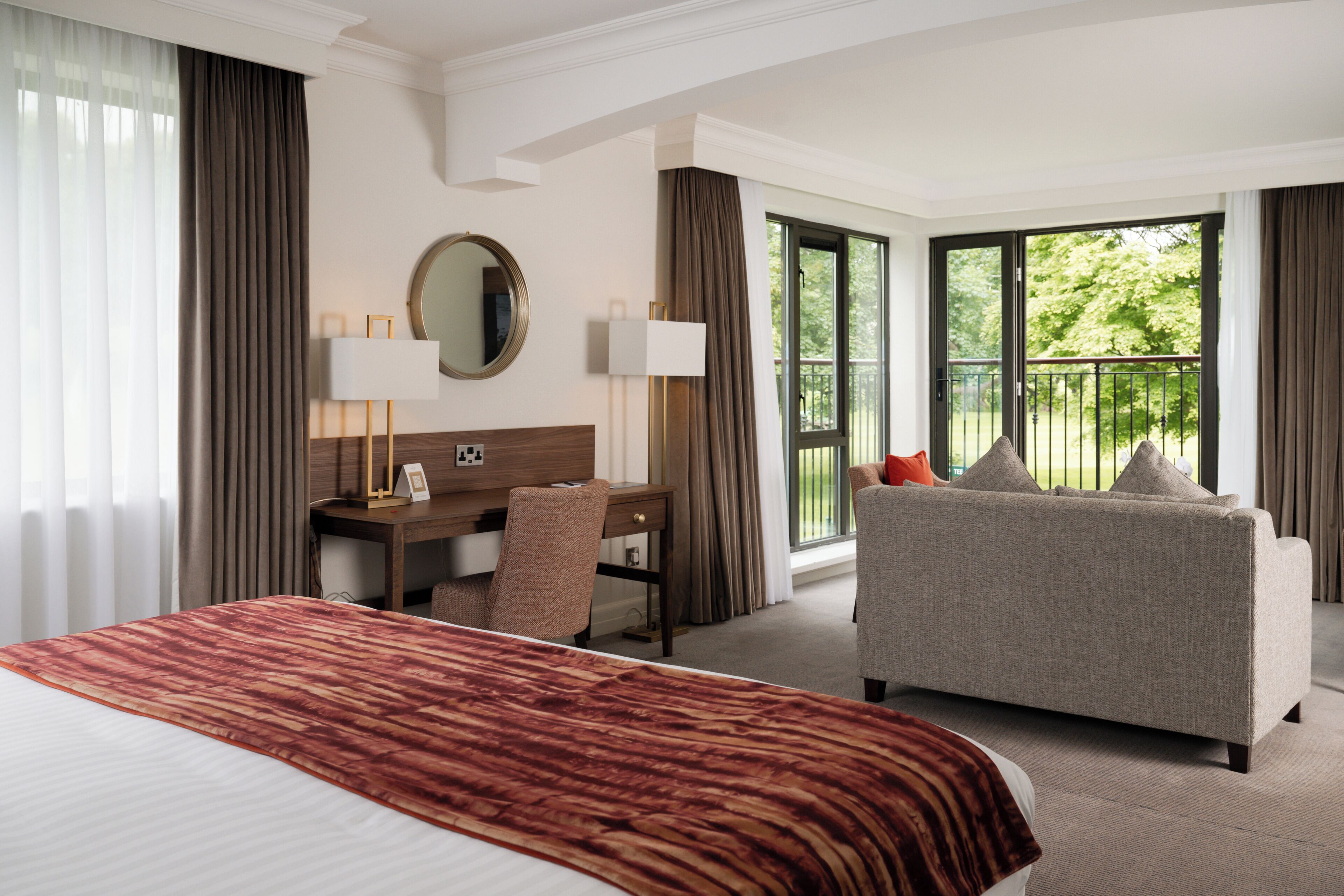 A hotel room with a large bed, a desk with a chair, a sofa, and a balcony with a view of greenery.