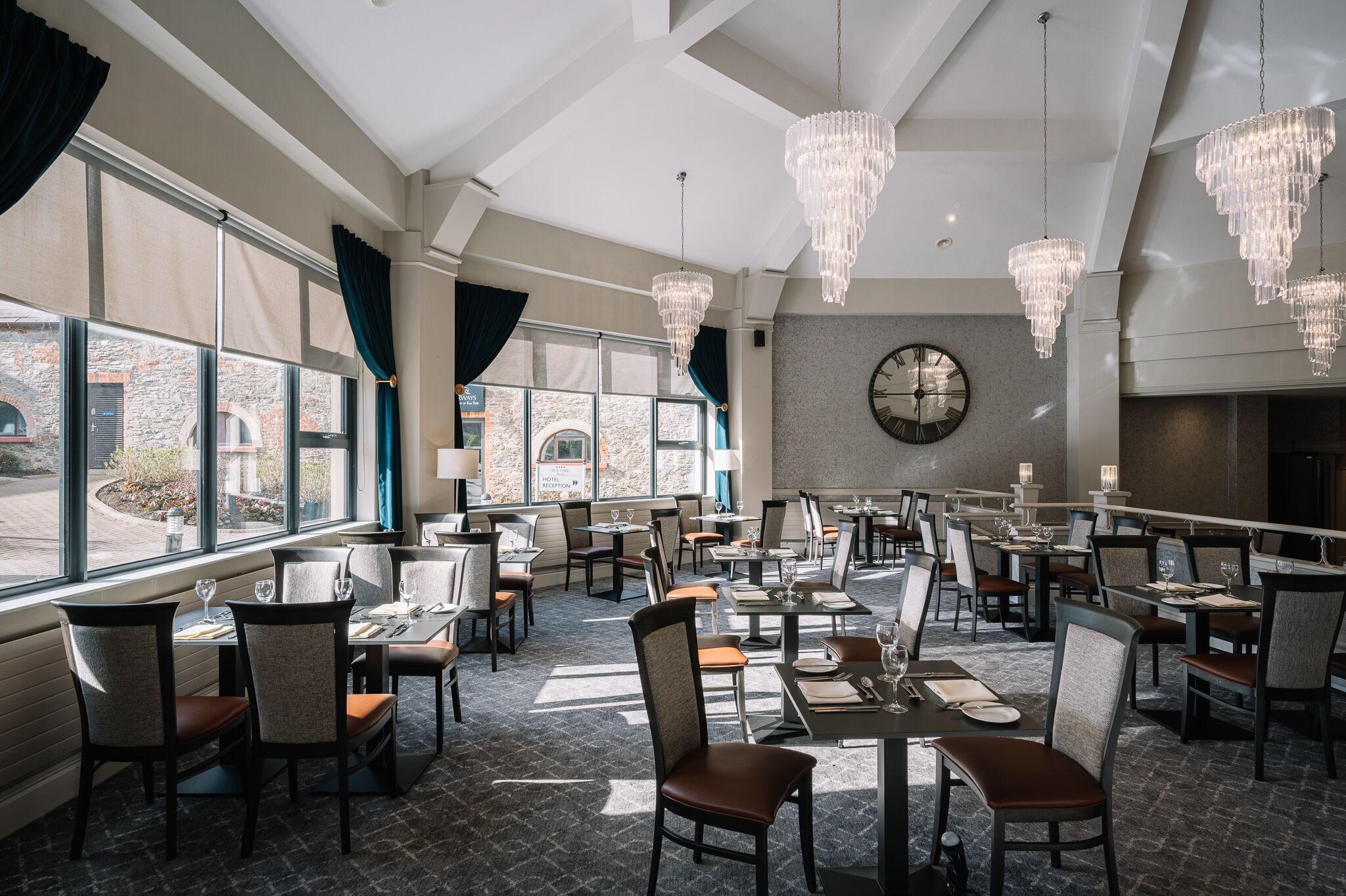A spacious dining area with multiple tables and chairs, large windows with curtains, and elegant chandeliers.