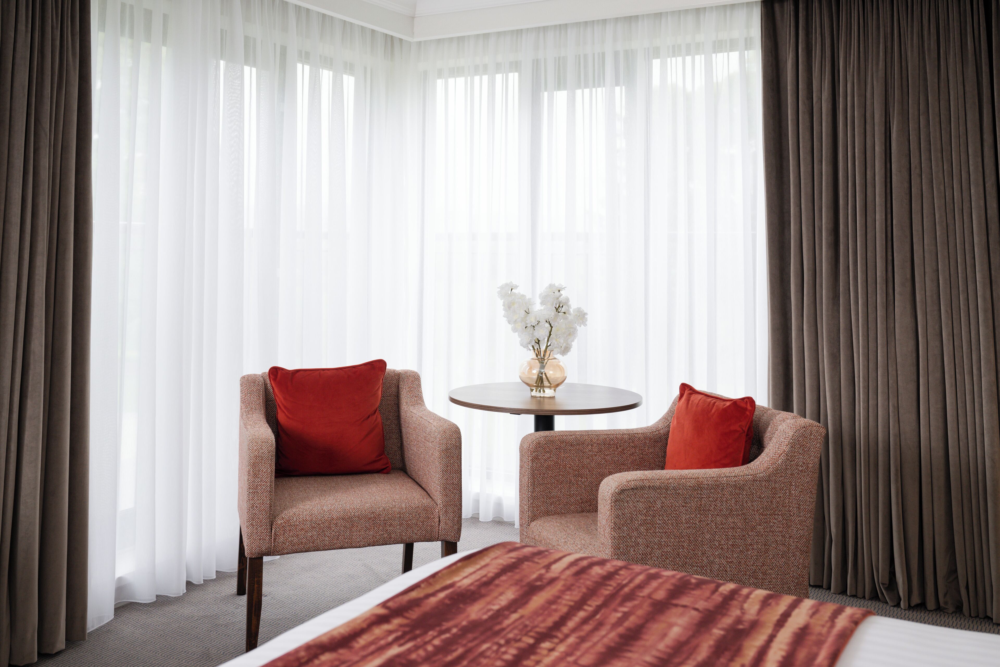 A hotel room with a bed, two armchairs, a round table with a vase of flowers, and sheer curtains.