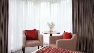 A hotel room with a bed, two armchairs, a round table with a vase of flowers, and sheer curtains.