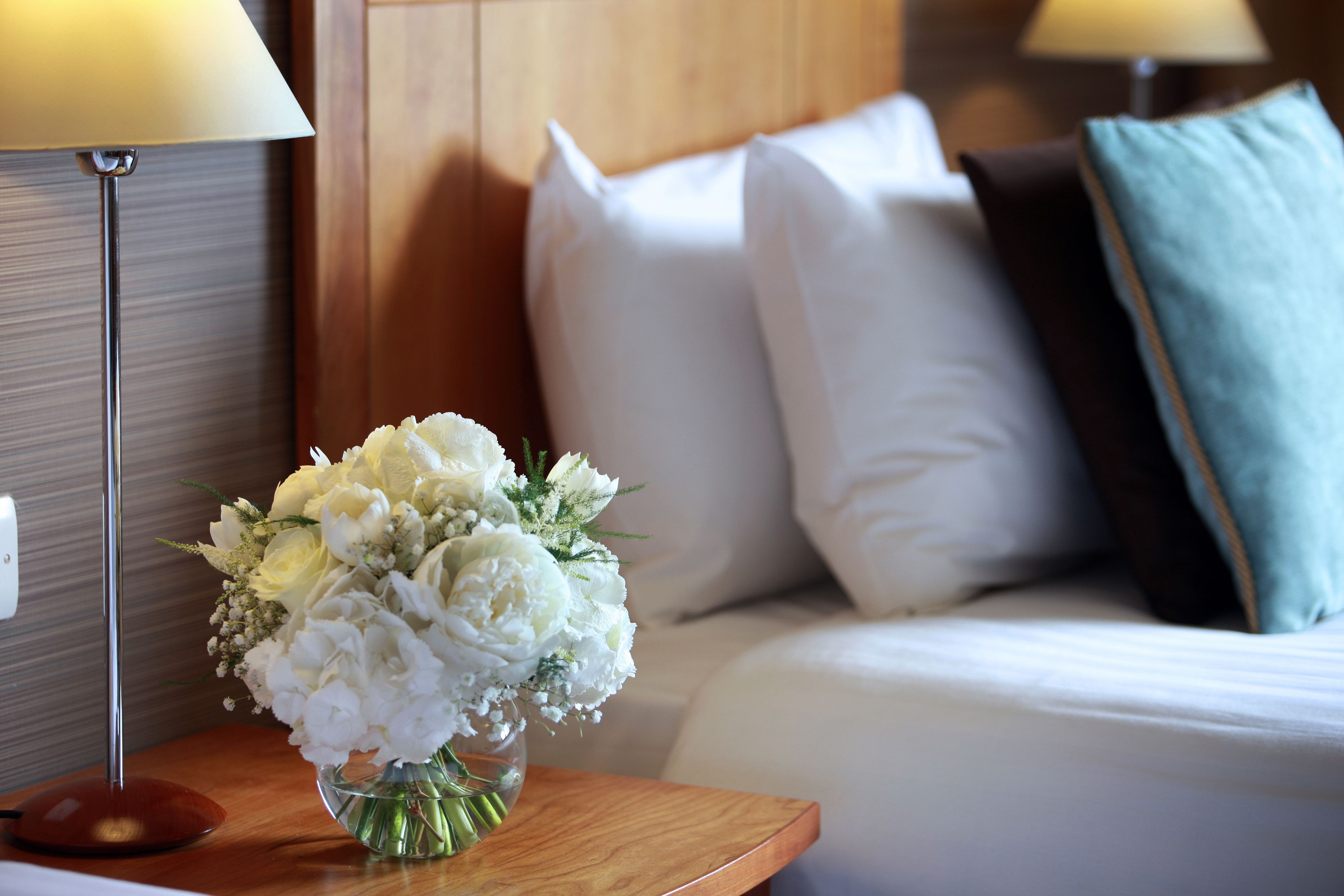 A hotel room with a bed, pillows, a bedside table with a lamp, and a vase of flowers.