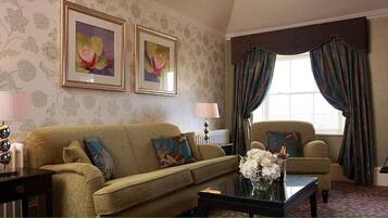 Una sala de estar con un sofá, sillones, una mesa de centro y un piano. Hay dos lámparas, un radiador y papel tapiz con motivos florales.