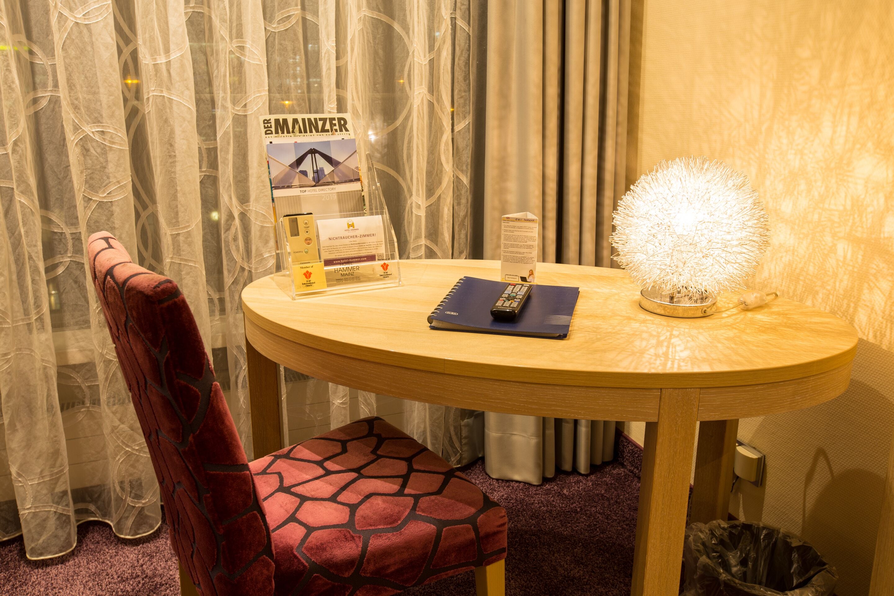 Una habitación de hotel con una mesa redonda de madera, una silla estampada de color rojo, un atril para folletos, un control remoto y una lámpara decorativa esférica.