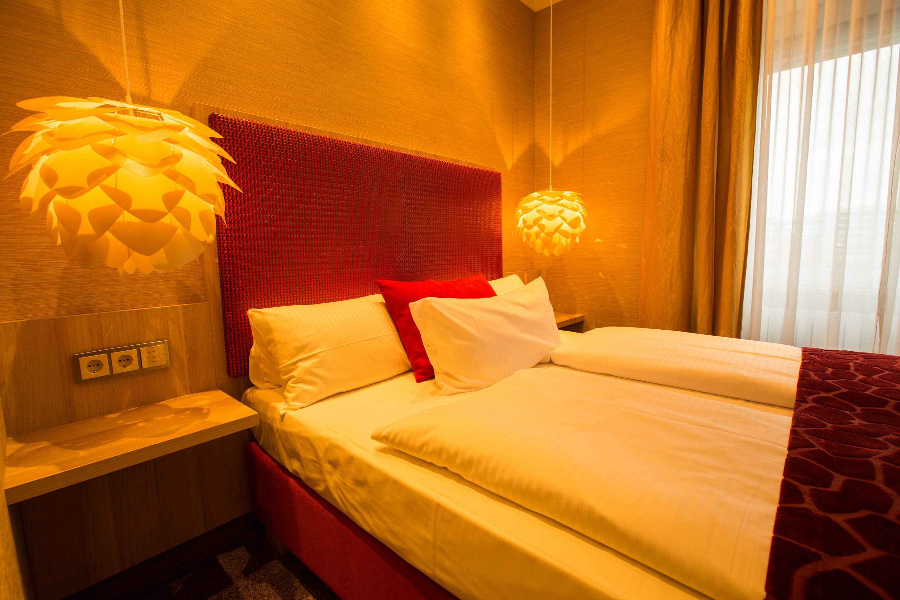 A hotel room with a bed, a nightstand, a red headboard, and two pendant lights.
