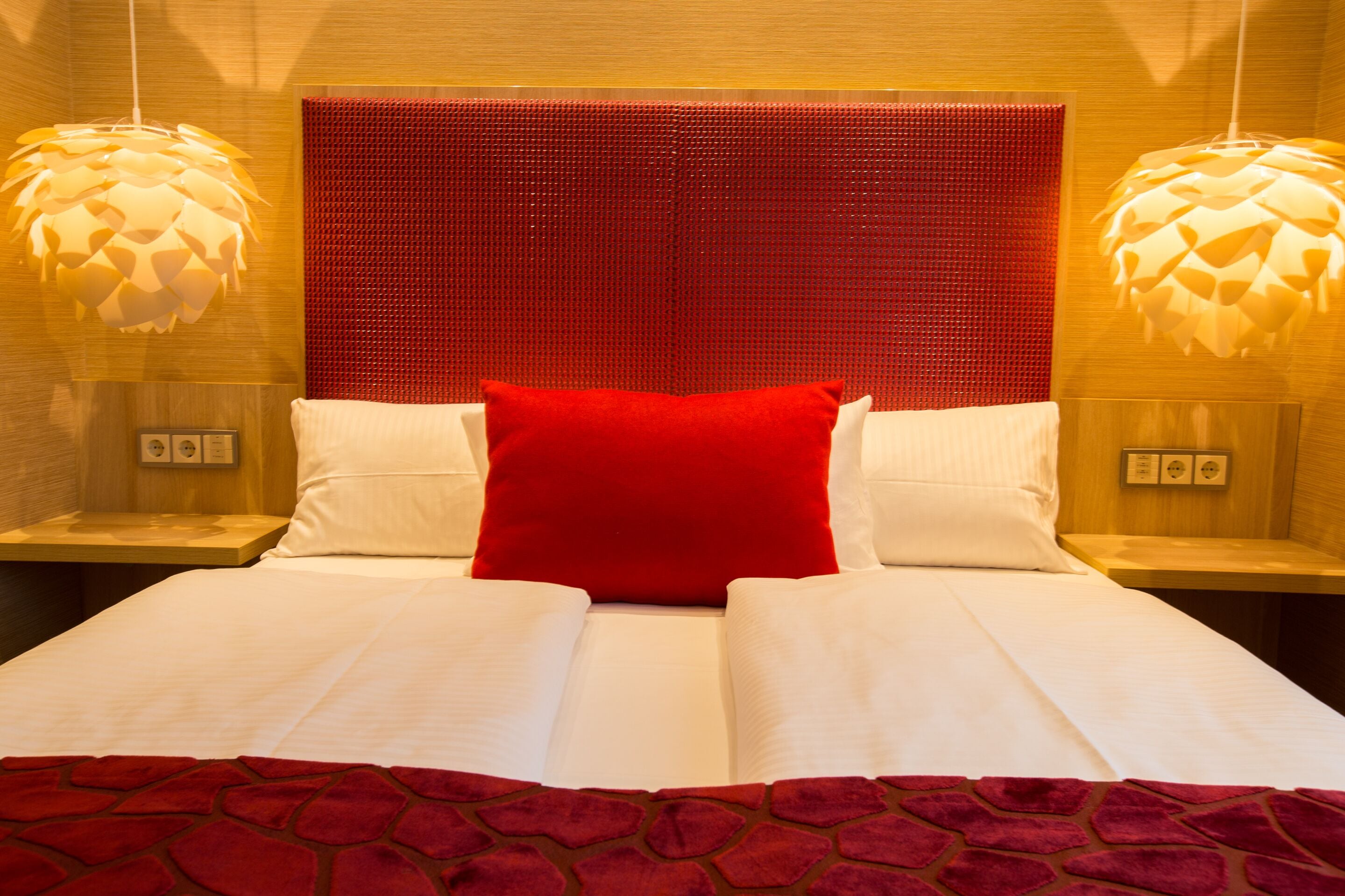A hotel room with a large bed, two bedside tables, a red headboard, and hanging lamps.