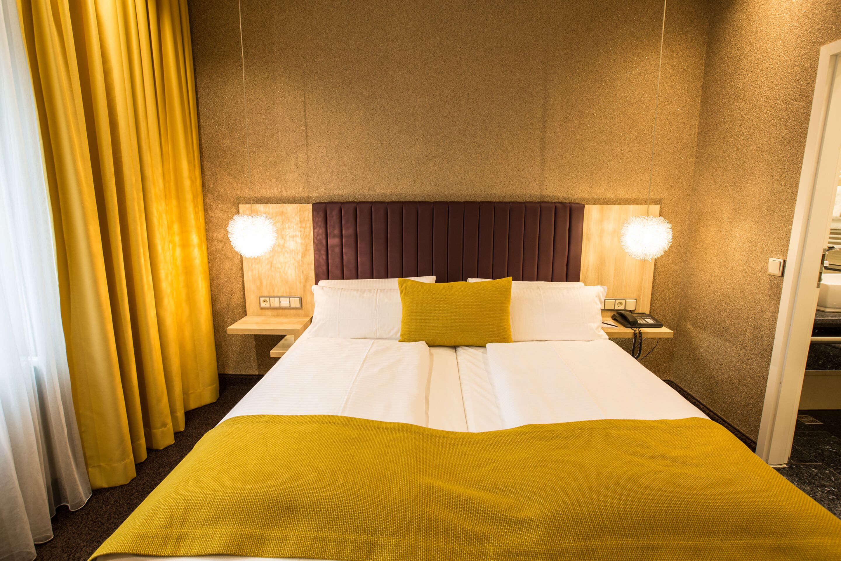 Quarto de hotel com uma cama grande, duas luminárias laterais, um painel de cabeceira de madeira e uma cortina amarela.