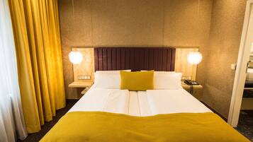 Quarto de hotel com uma cama grande, duas luminárias laterais, um painel de cabeceira de madeira e uma cortina amarela.