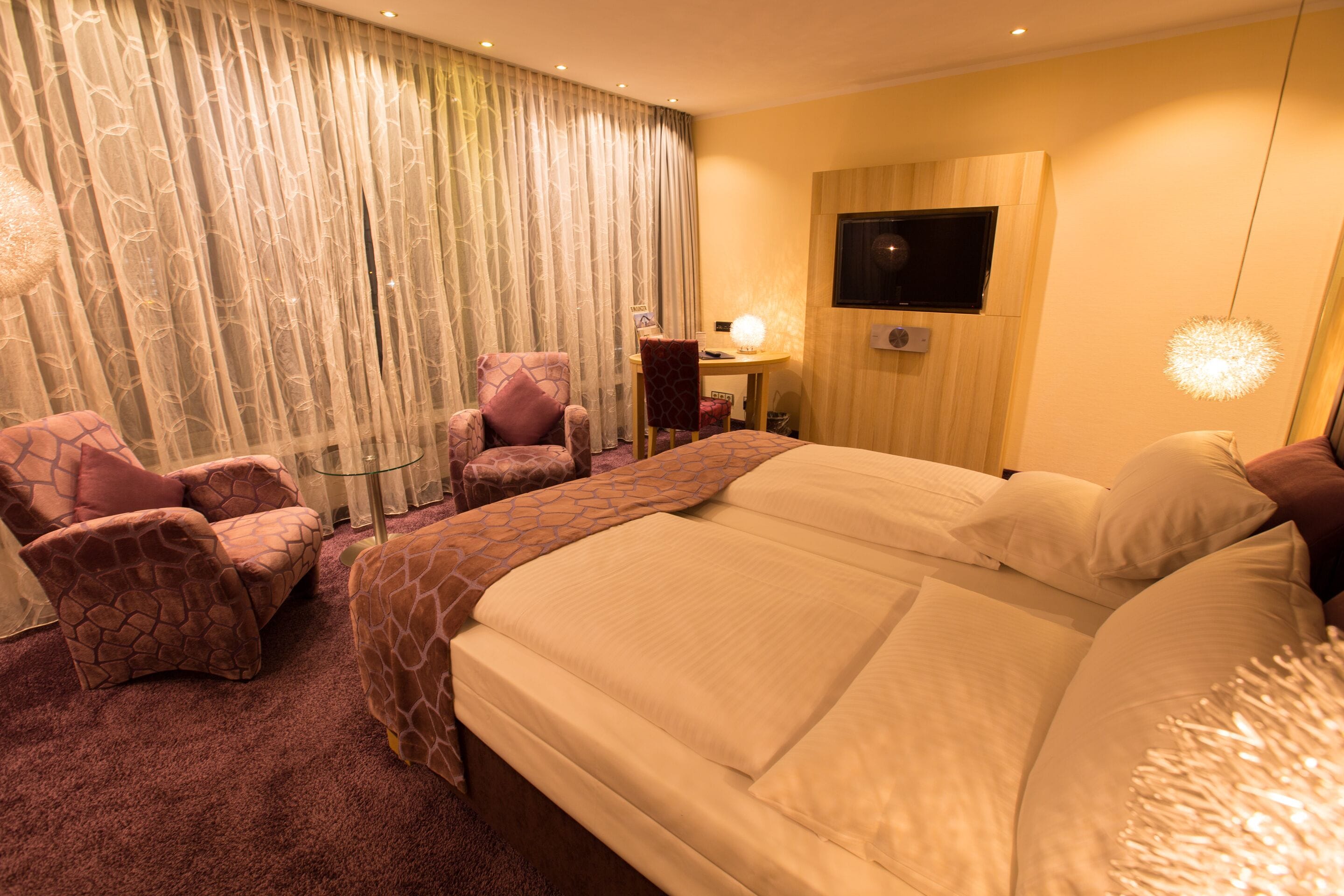 A hotel room with a large bed, two armchairs, a desk with a lamp, and a TV on the wall.