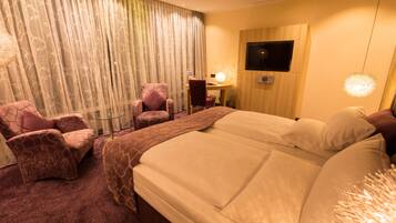 A hotel room with a large bed, two armchairs, a desk with a lamp, and a TV on the wall.