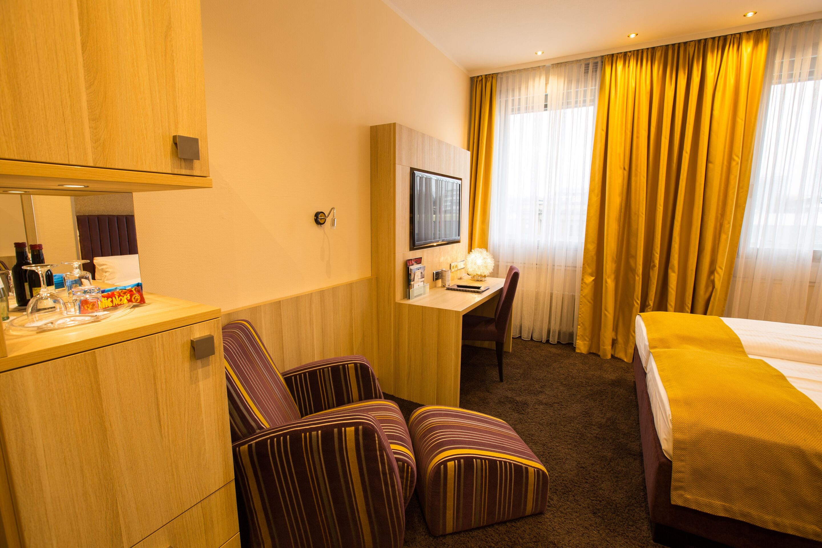 A hotel room with a bed, a desk with a chair, a television, and a minibar.