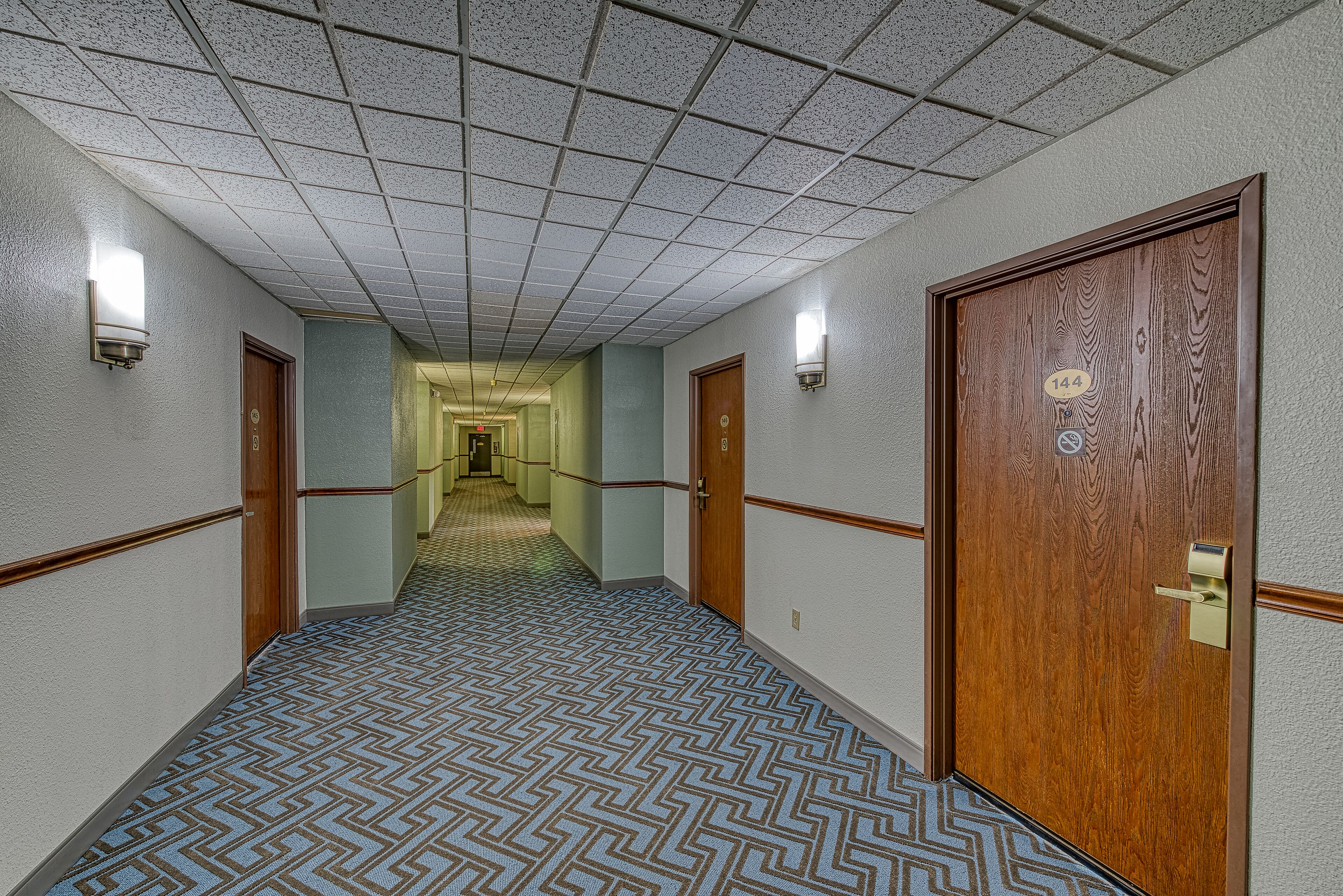 Um corredor de hotel com carpete estampado, portas de madeira e teto em azulejos.
